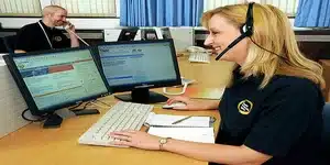 Een vrouw met een headset zit achter een computer en lijkt ondersteuning te bieden, terwijl op de achtergrond een man aan de telefoon is, wat wijst op een kantooromgeving voor klantenservice.