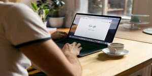 Een persoon zit aan een houten tafel en gebruikt een laptop die Google als startpagina weergeeft. Er staat een kopje koffie naast de laptop.