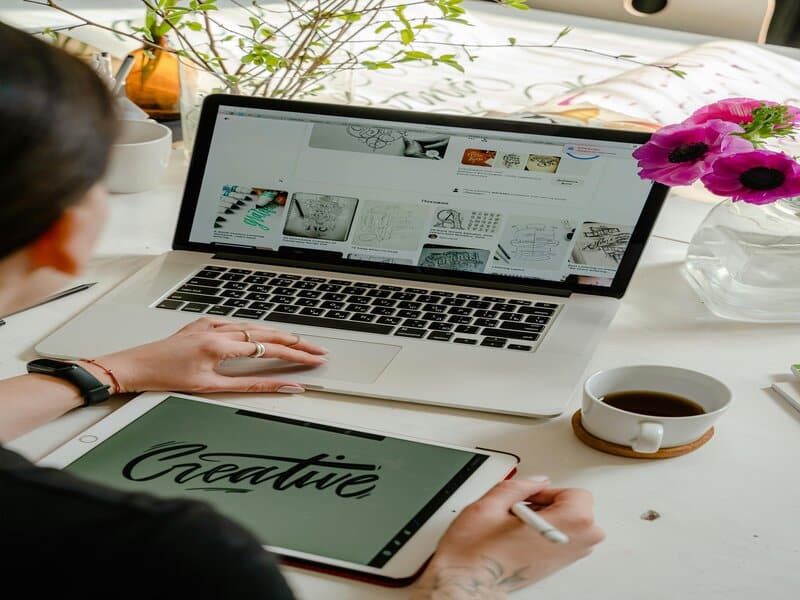 Foto van een persoon die werkt aan een laptop met inspirerende afbeeldingen en een tablet met het woord 'Creative', een typisch scenario in de wereld van creative en digital agencies.