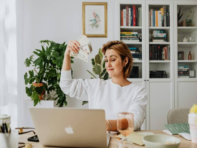 Foto van een vrouw die geldbiljetten vasthoudt terwijl ze naar een laptop kijkt, wat wijst op de winstgevendheid en het inkomen in de sector van marketing.