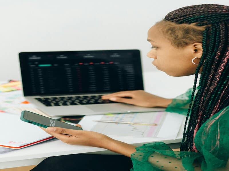 Foto van een jonge professional die aan een bureau zit, met een laptop en een smartphone in de hand, geconcentreerd op schermen met grafieken, wat representatief is voor data-analyse in online marketing in NL.